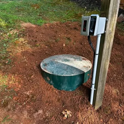 A sewage pump basin set in red clay beside a post carrying its control box and alarm